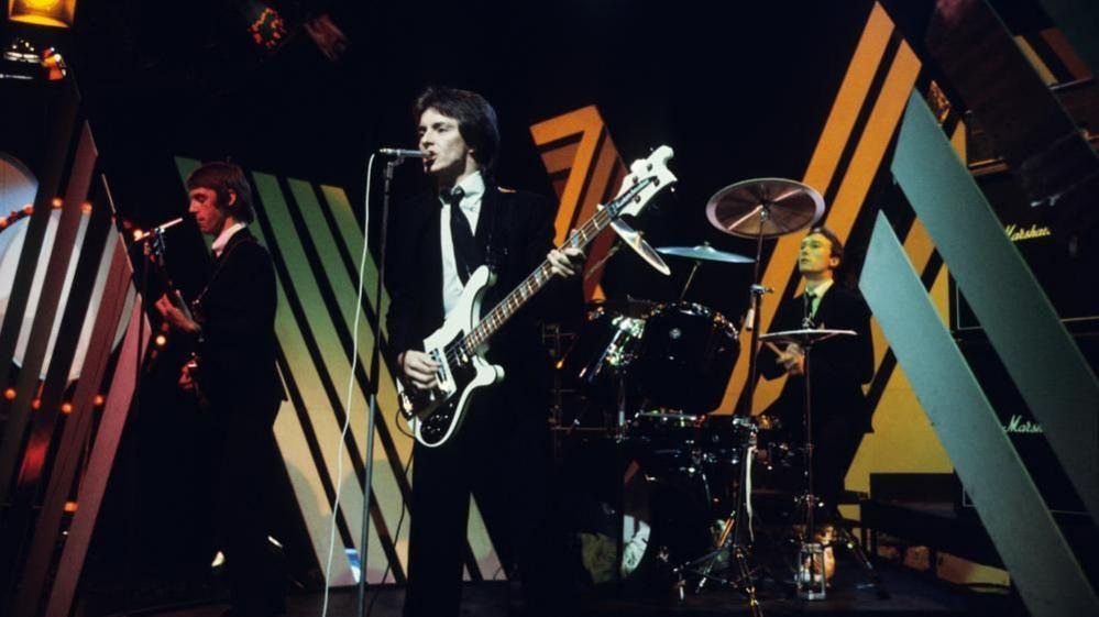 Rick Buckler is pictured on drums with The Jam bandmates Paul Weller (left) and Bruce Foxton (centre) performing in 1977 on the BBC's Top of the Pops show. They are dressed in black suits, white shirts and black ties against a BBC stage set with Marshall sound kit in the background.