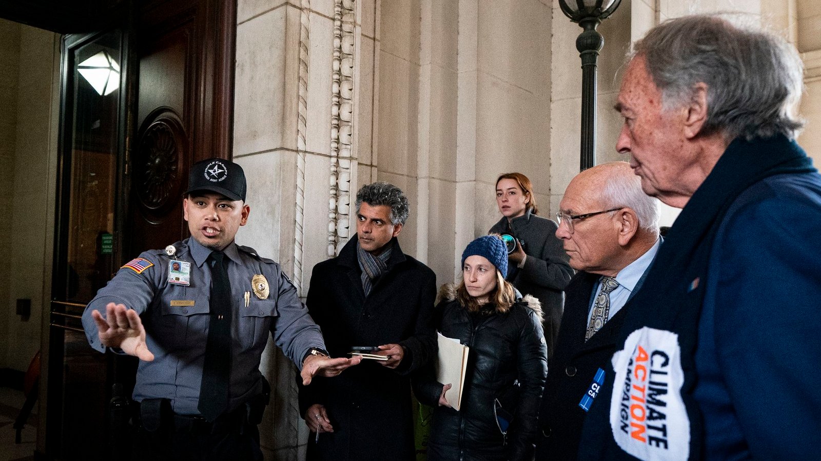 A security guard in a gray uniform gestures to a group of lawmakers and protesters outside a marble building.