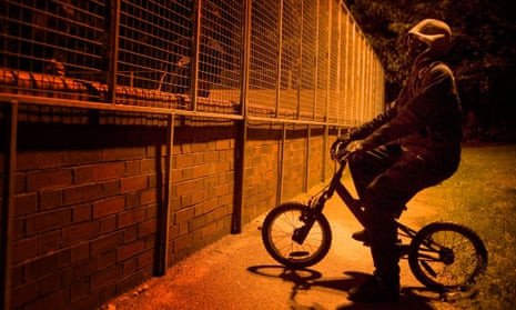 Anonymous teenager on a BMX-type bicycle talks to two shadowy figures through a wire fence; it is dark and his face is covered with a scarf and hood.