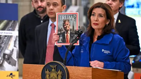 Getty Images New York Governor Kathy Hochul holds up a photo of a fake magazine cover showing Donald Trump wearing a king's crown with the words, "long live the king" during a news conference in a subway station.