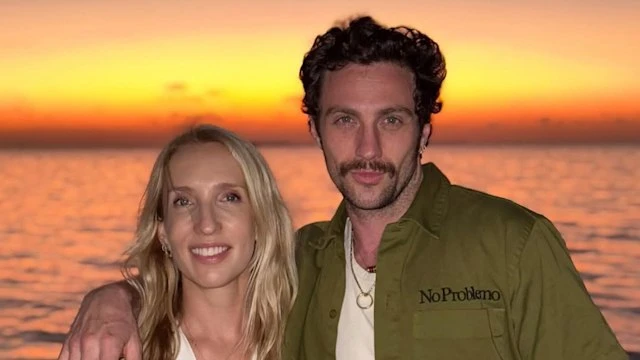 Aaron Taylor-Johnson puts his arm around his wife Sam while posing in front of a sunset