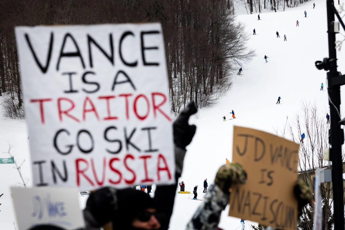 Protesters holding signs criticize JD Vance, referring to him as a traitor, at a snowy ski resort with skiers in the background.