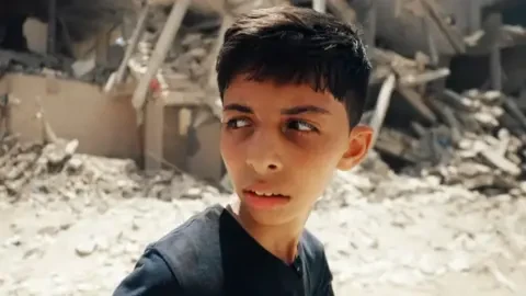 BBC/AMJAD AL FAYOUMI/ HOYO FILMS A child in Gaza with rubble behind him