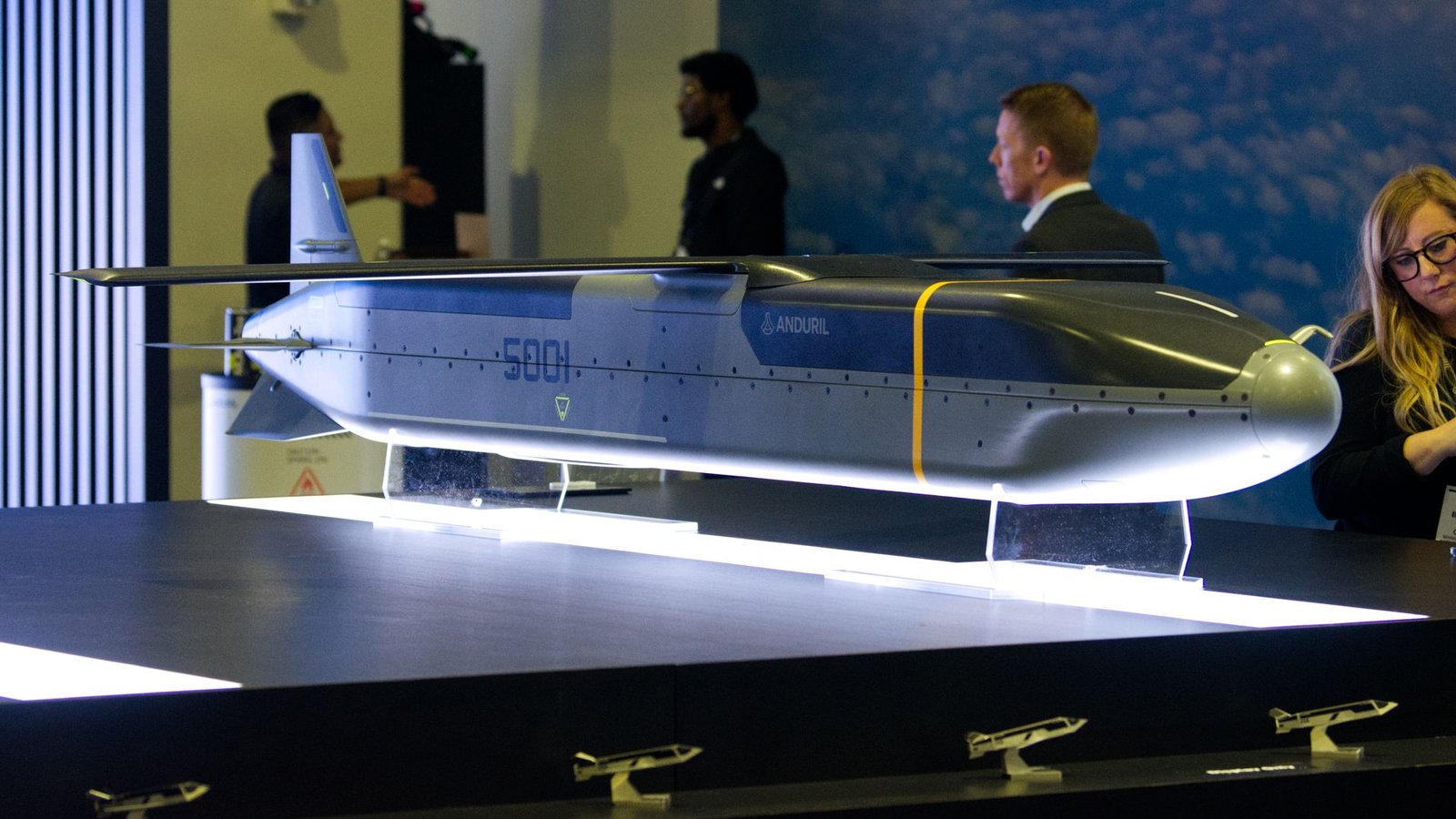 An Anduril Industries Barracuda cruise missile is perched at the companies booth at a defense conference. Four people are seen behind it.