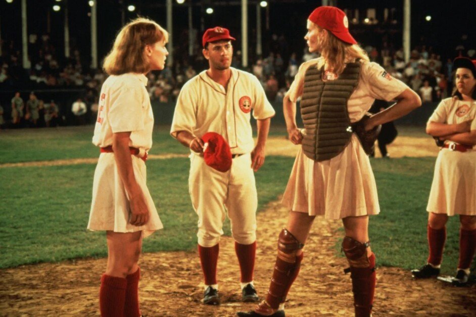 Two female baseball players in uniforms have a discussion on the field with their male coach. One of the female players is a catcher, wearing protective gear. It appears to be nighttime with spectators in the background.