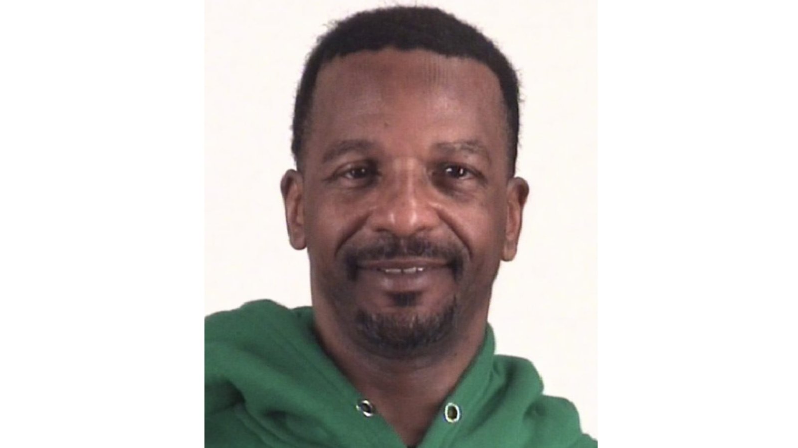 A man wearing a green hoodie smiles at the camera against a plain background.