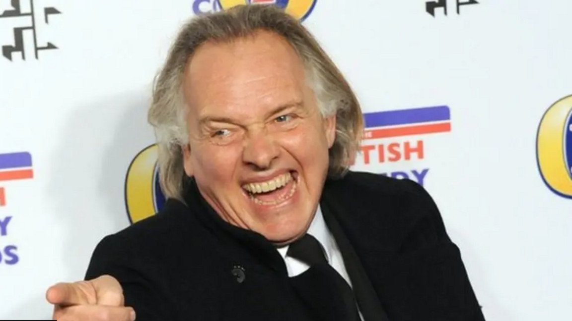 Rik Mayall in front of a British Comedy Awards backdrop, laughing and pointing