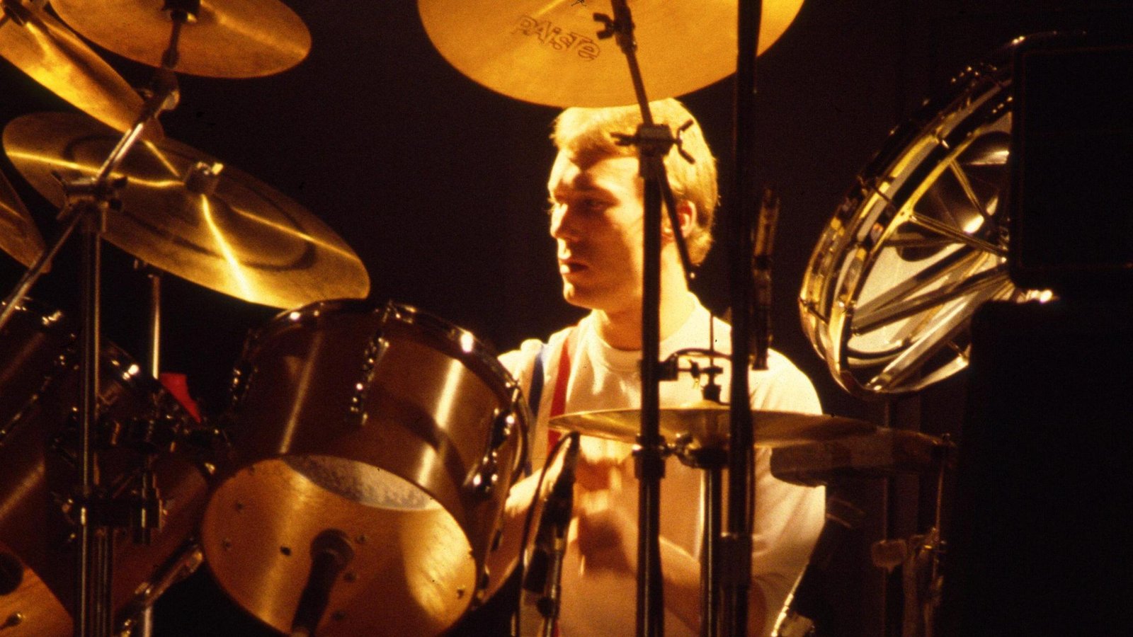 Rick Buckler behind a drum kit on stage, against a black background. He and his drums are lit up with an orange-sepia colour.