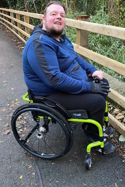 Kieren is sitting in a florescent green wheelchair in a country park. He is a white man, sporting a beard and hair swept backwards. Kieren is looking at the camera with a smile.