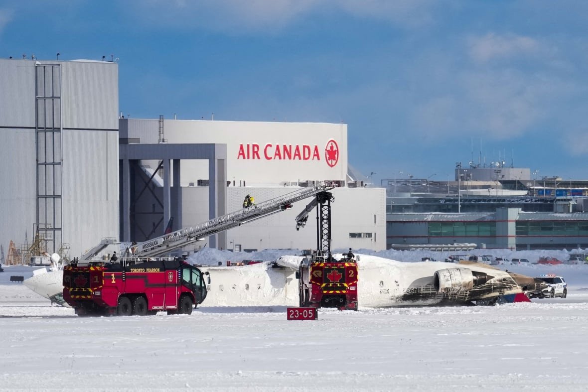 On a snowy runway at Pearson airport, emergency vehicles are parked beside an overturned plane. An Air Canada hangar is visible in the background. It's a blue sky day.