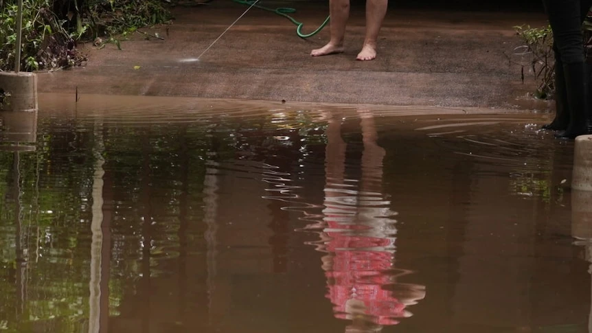 A woman is reflected in a large puddle as she hoses a driveway.