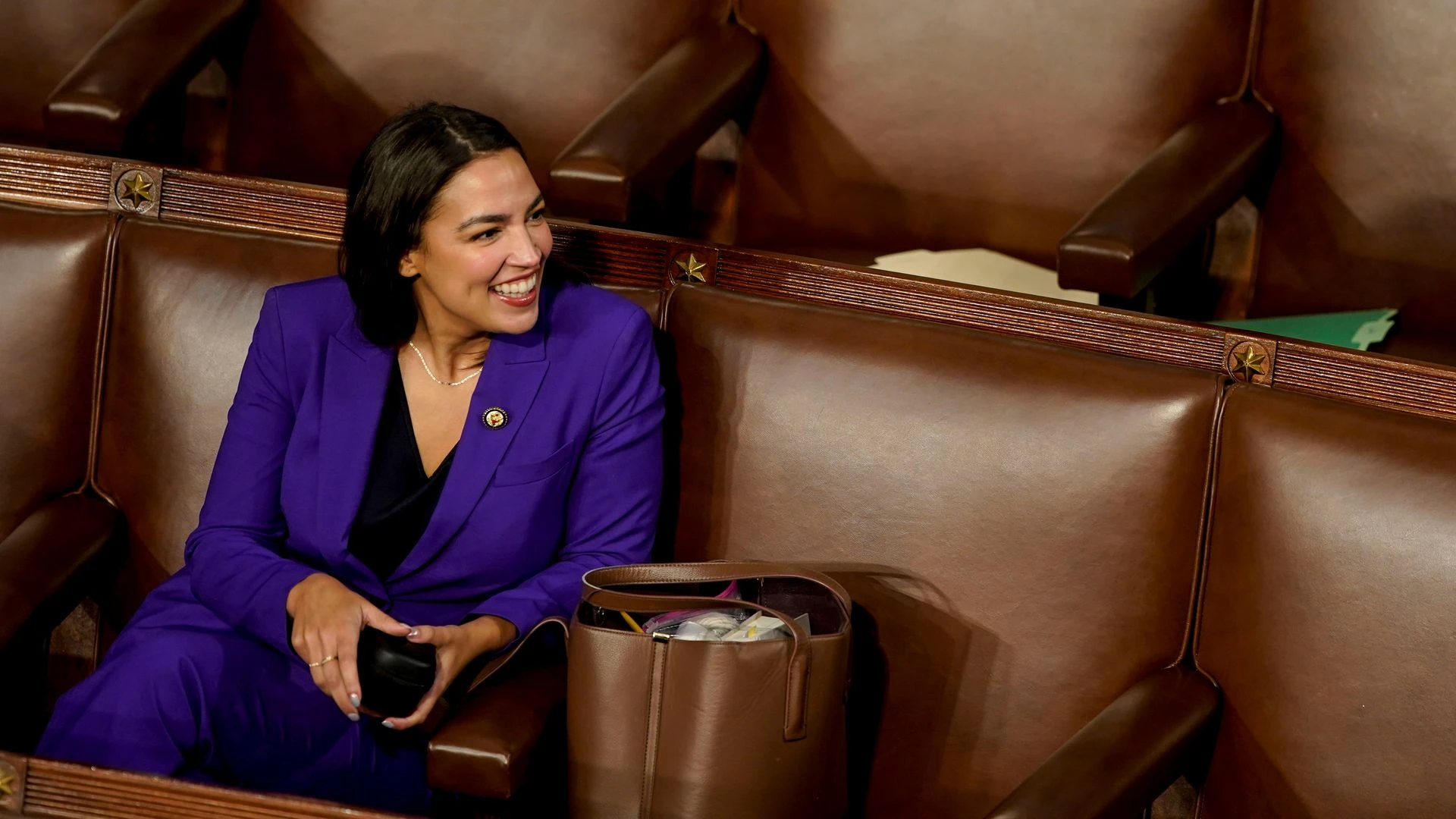 Rep. Alexandria Ocasio-Cortez, wearing a purple suit and sitting in a brown leather chair in the House chamber.