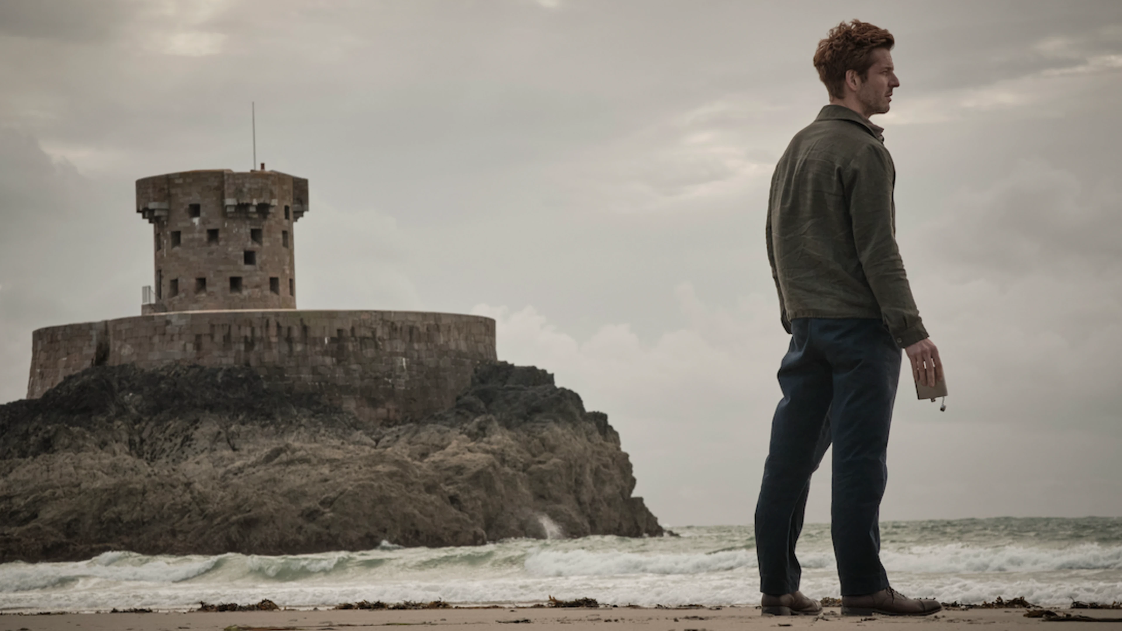 Damien Molony is holding an empty alcohol flask looking out onto the ocean. He's standing on St Ouen's Bay with La Rocco tower and a moody, grey sky behind him.